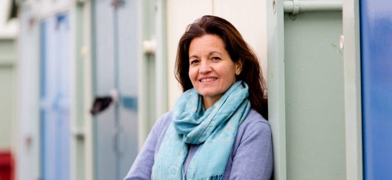 A picture of me leaning against a hut on Brighton seafront wearing a blue jumper and blue scarf. smiling to the camera