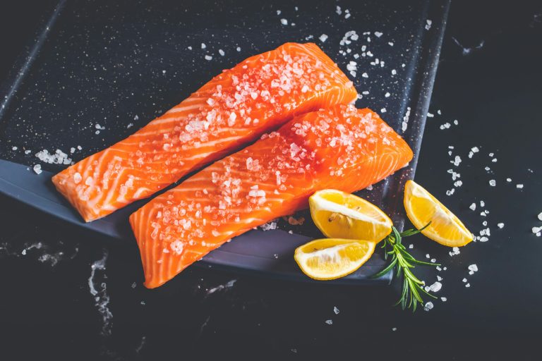 Picture of raw wild salmon fillets with sea salt and lemons and herbs next to the fillets on a slate