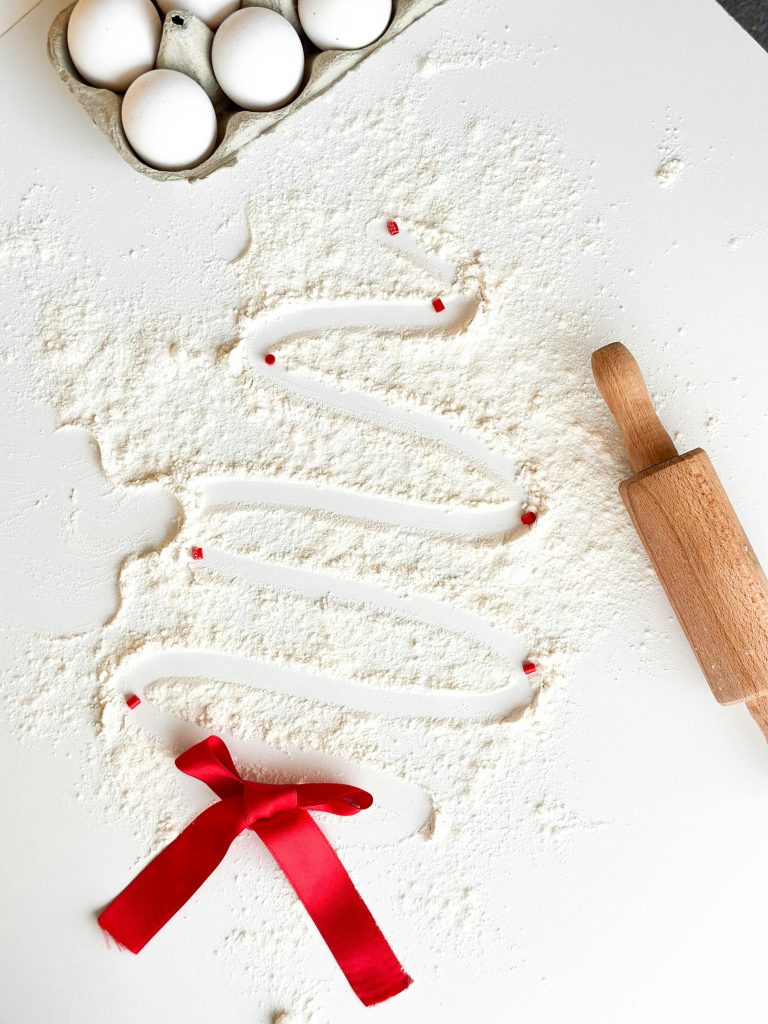 A xmas tree drawn with a finger out of white flour on a counter top wiht a red ribbon at the bottom and a rolling pin and eggs next to it